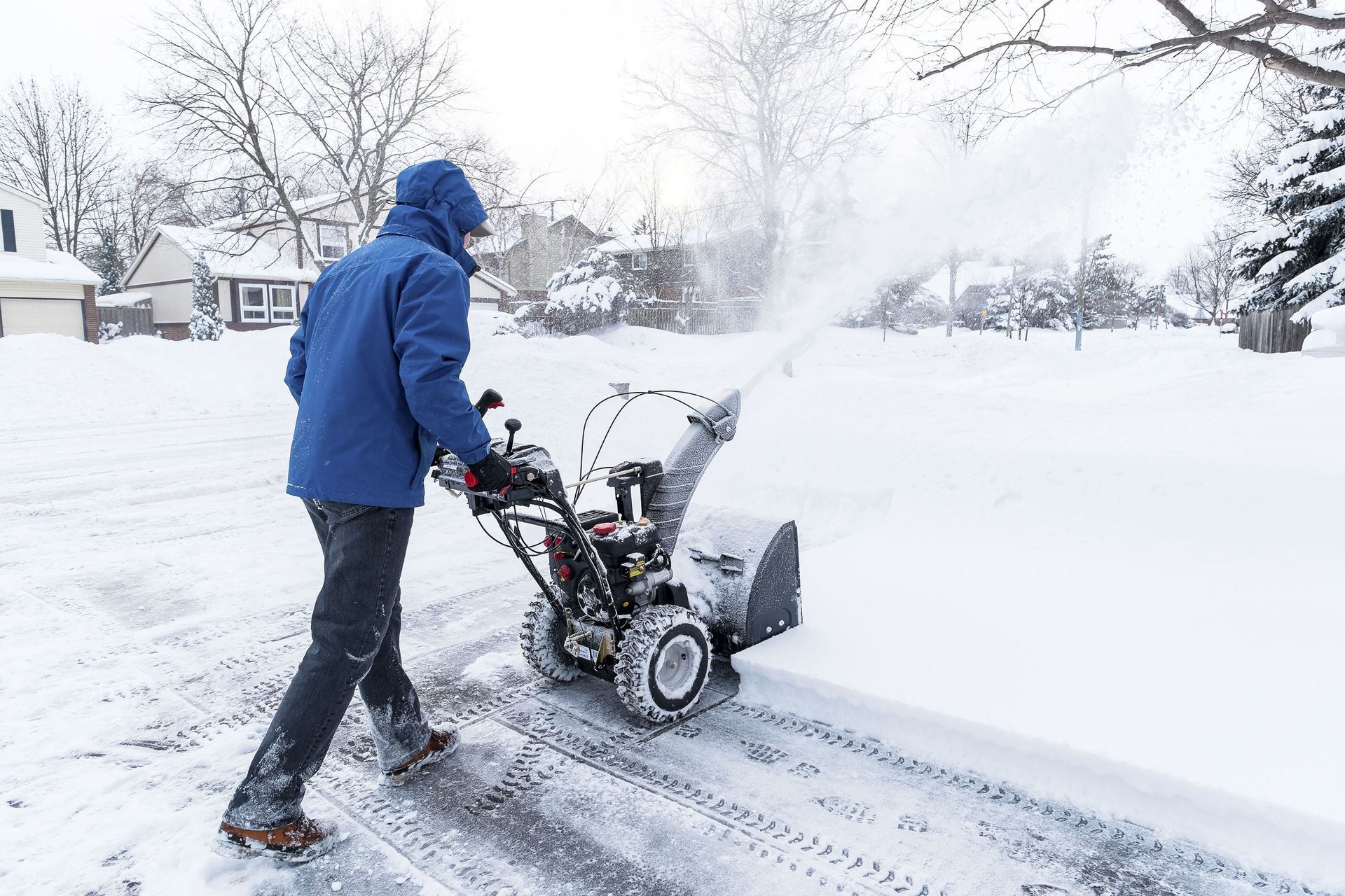 Person in blue jacket using a snowblower to clear a snowy driveway.