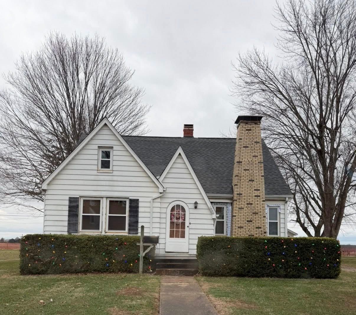 White house with dark roof and stone chimney, framed by hedges and bare trees on a gray day.