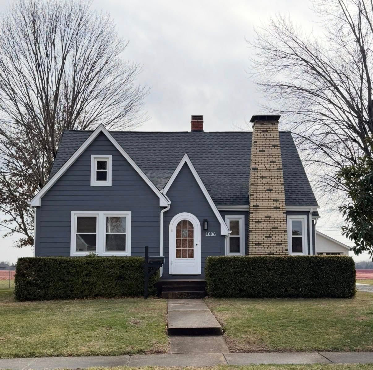 Small blue-gray house with white trim, a tall stone chimney, and bare trees in front