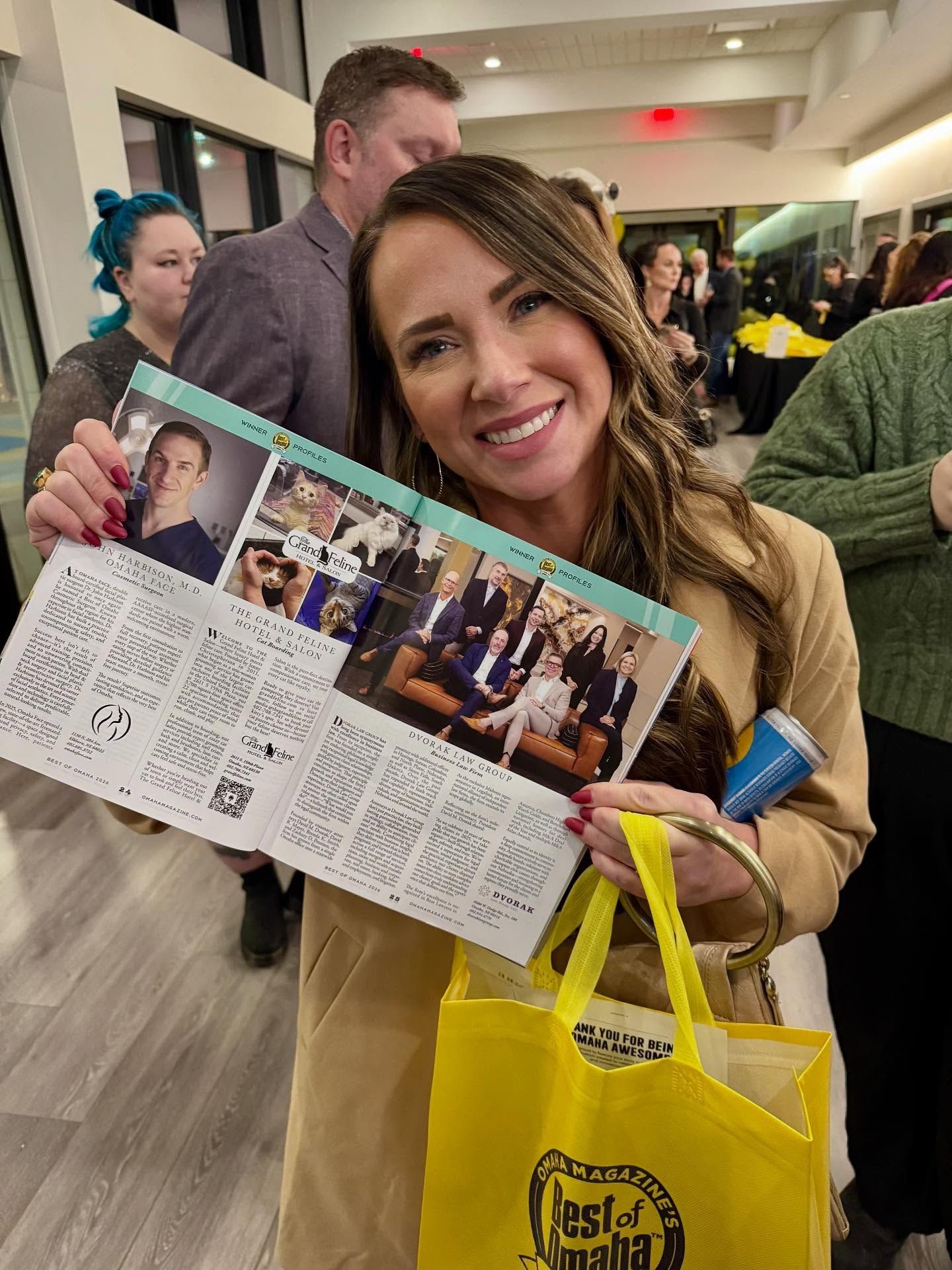 Woman smiles, holding a printed program & yellow tote bag. Other people in background. Beige coat.