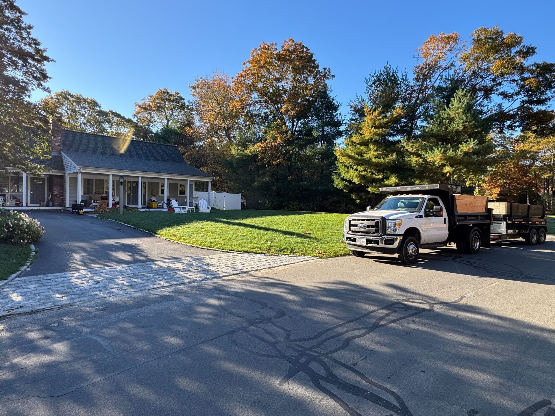 White pickup truck with a trailer parked in front of a house with fall foliage, on a sunny day.