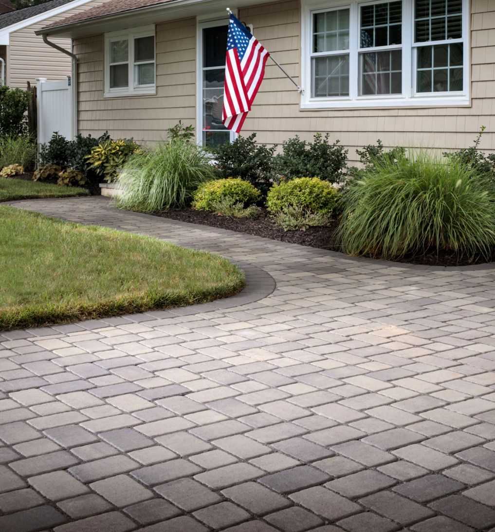 Brick driveway leading to a beige house with an American flag.