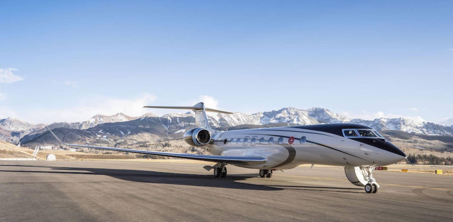 Private jet on an airport runway with snow-capped mountains and blue sky in the background.
