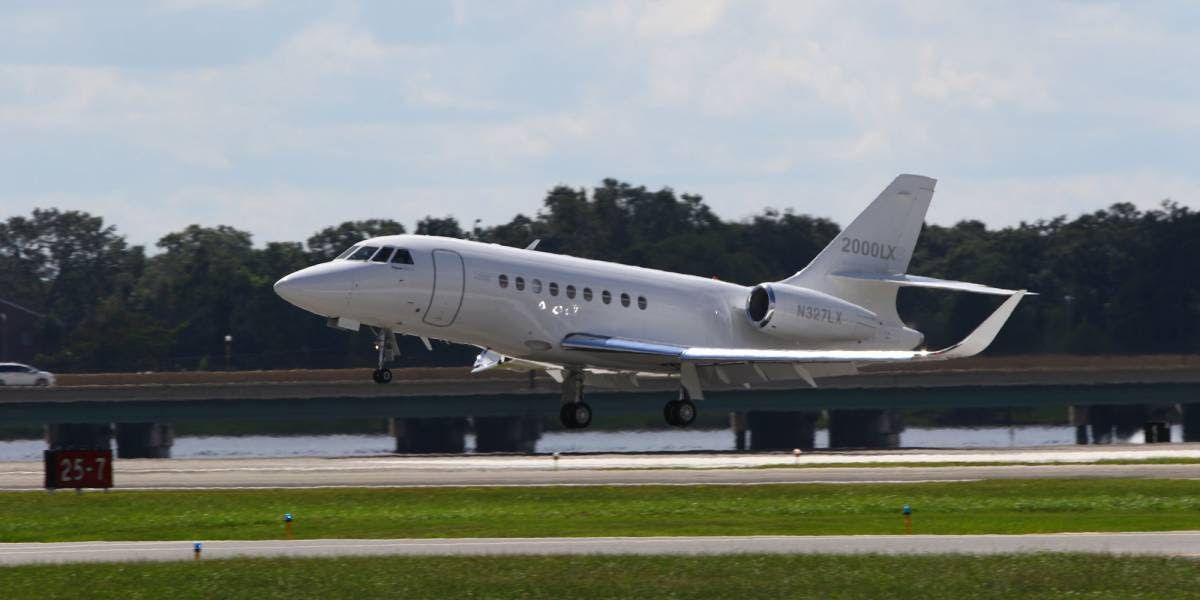 A white private jet takes off from a runway. Green grass and trees are in the background.