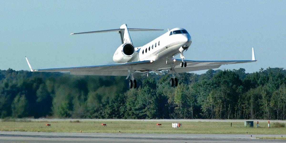 A white private jet lands on a runway, with green trees in the background under a blue sky.
