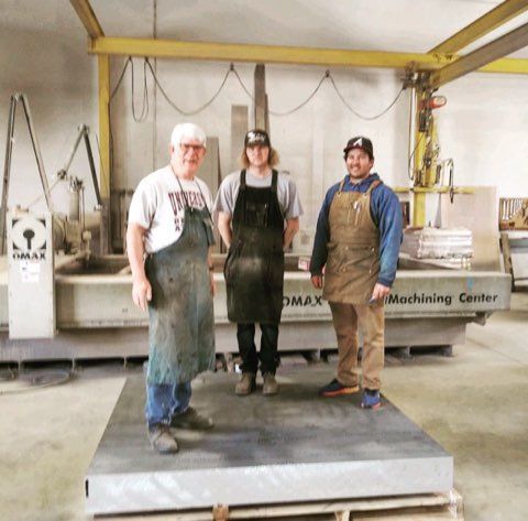 Three people stand on a large stone block in a workshop, near a waterjet machine.