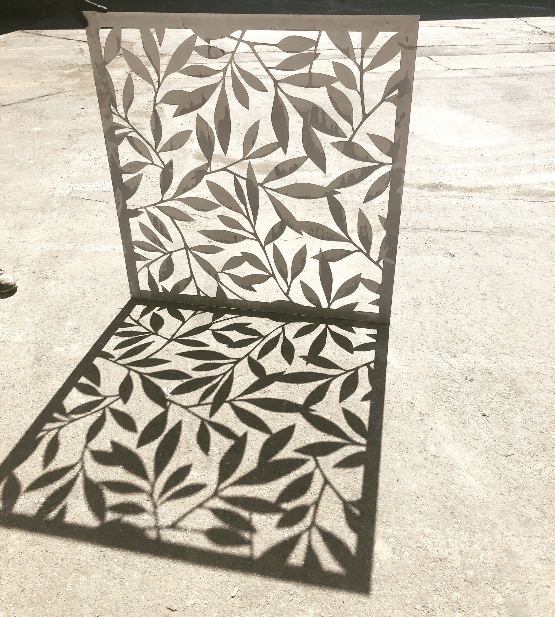 Metal panel with leaf pattern casting a leafy shadow on pavement.