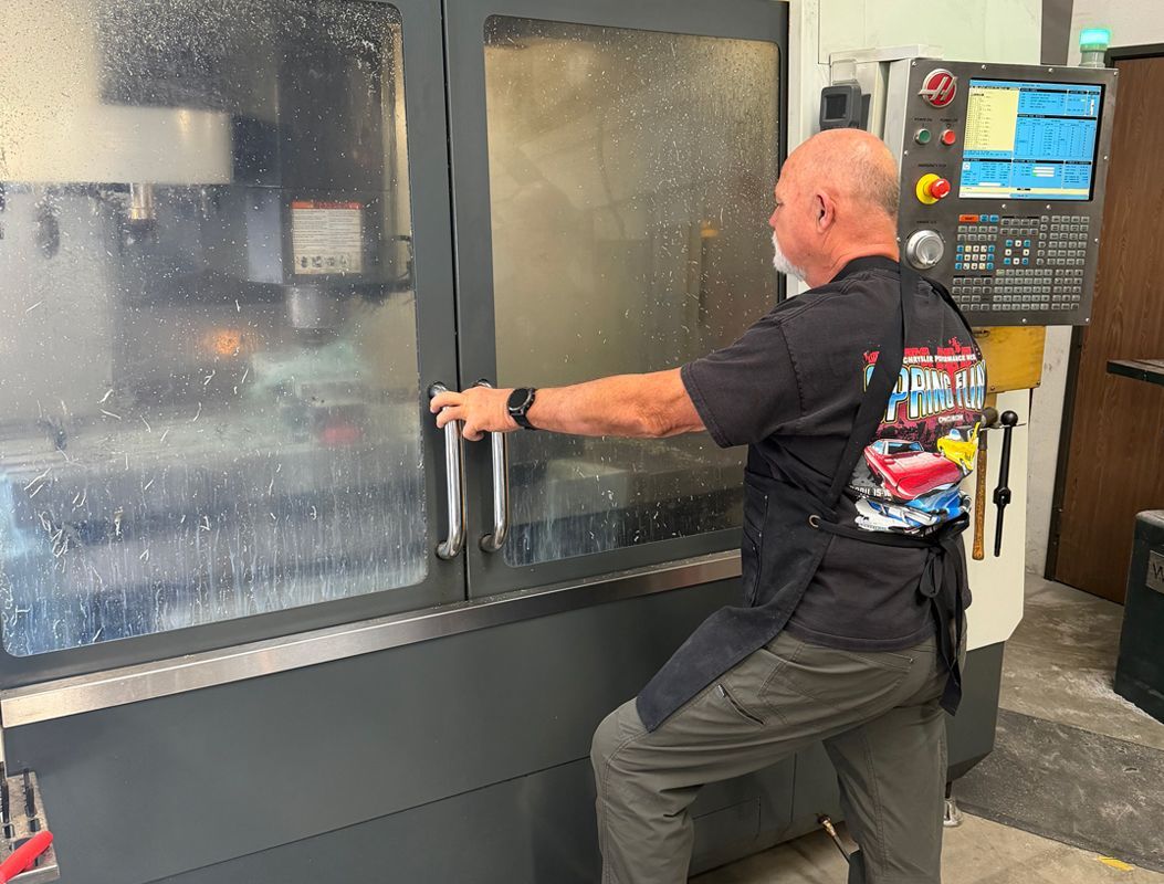 A person operating a CNC machine in a workshop, wearing a black shirt beside a control panel.
