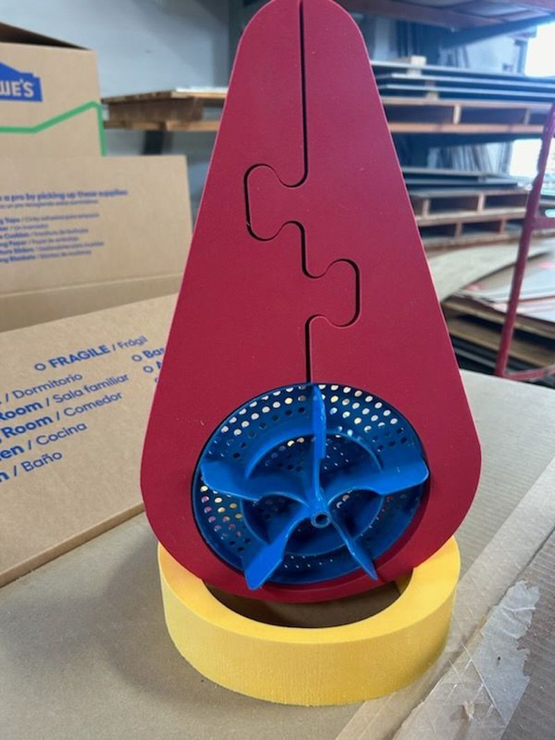 Red teardrop-shaped wooden puzzle with a blue circular insert on a yellow base in a workshop.