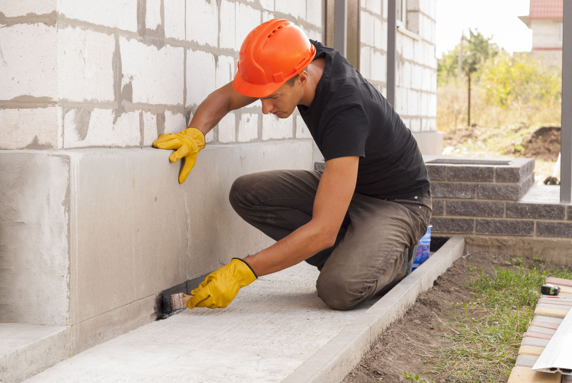 Construction worker applying sealant to a foundation, wearing an orange hard hat and yellow gloves.
