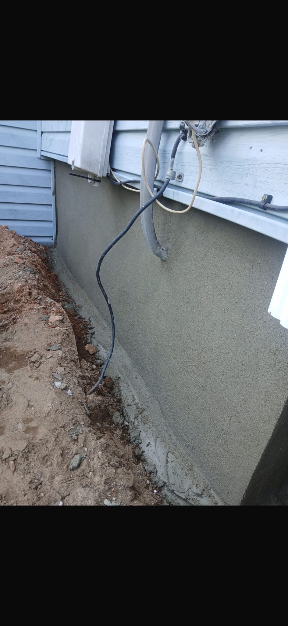 A freshly patched, textured concrete foundation wall along the side of a house with exposed soil and utility wiring.