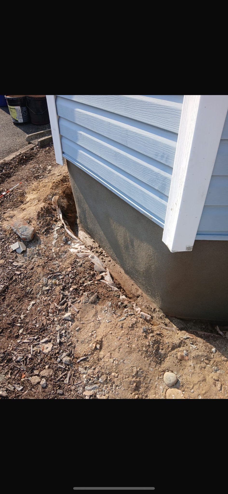 A trench excavated alongside the concrete foundation of a house with light blue siding.