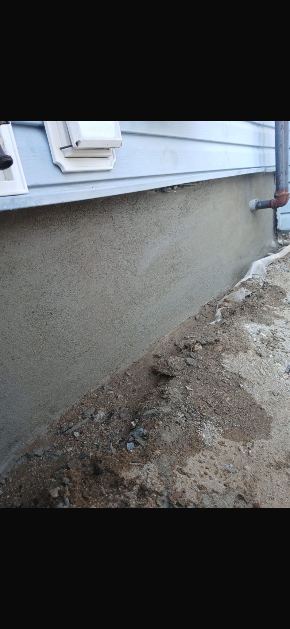A section of a house foundation with a grey, textured parge coat finish, situated above a dirt and gravel ground surface.
