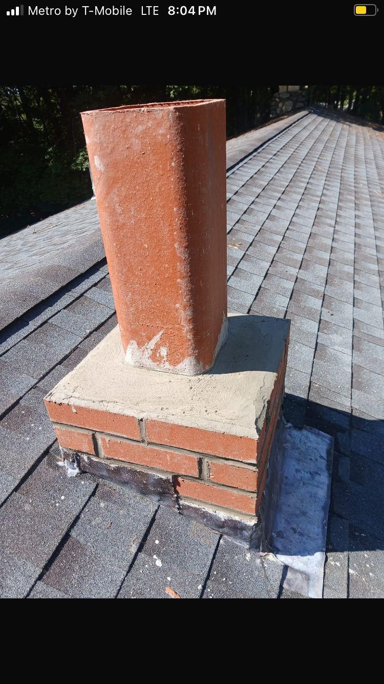 A red brick chimney stack on a residential shingled roof with a concrete base and sealant visible around the edges.