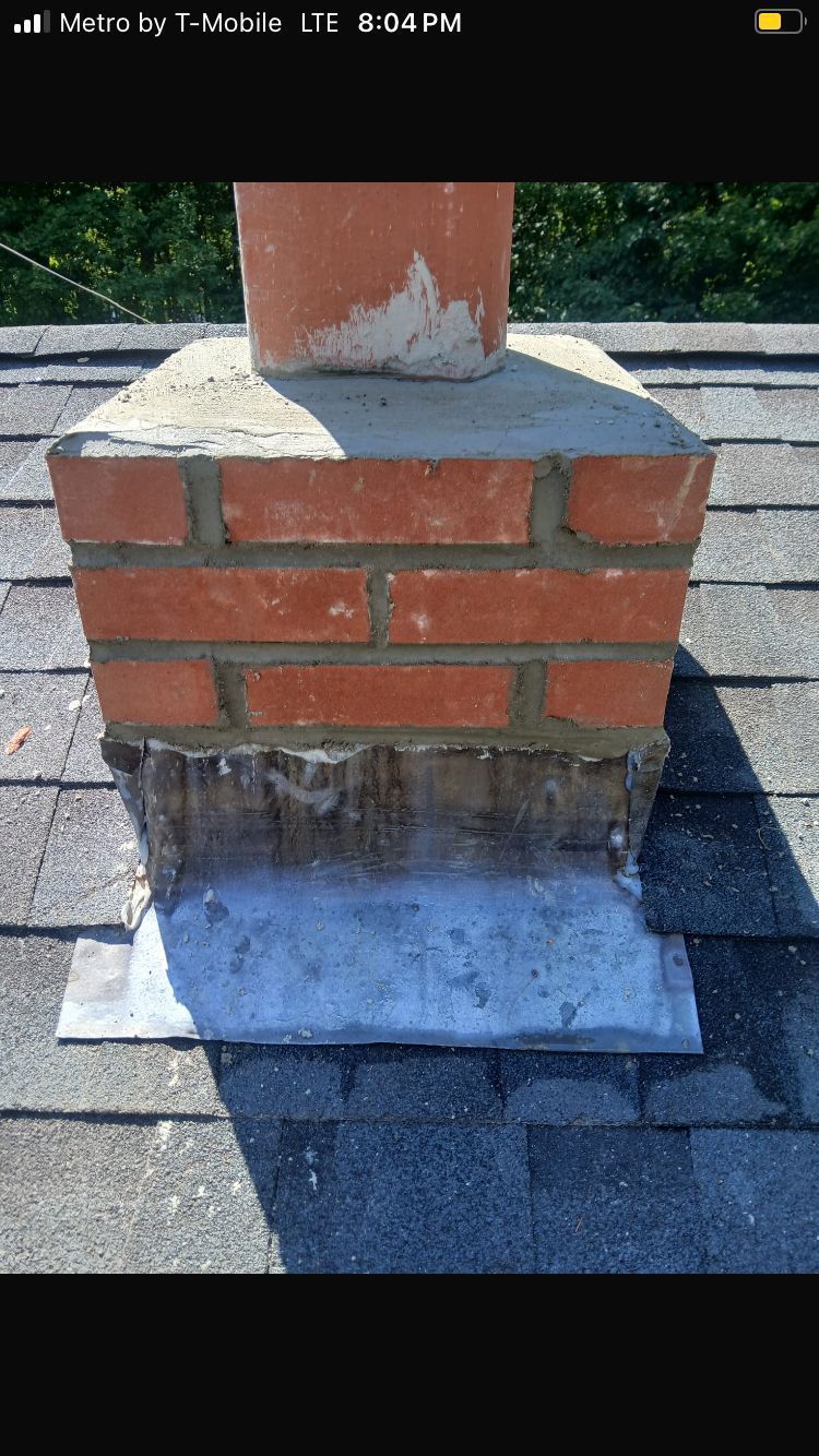 A brick chimney base on a shingled roof, featuring a freshly applied concrete mortar crown and metal flashing at the base.
