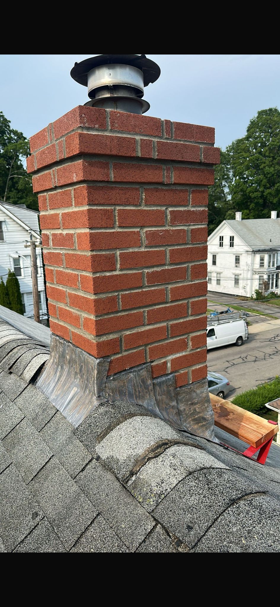 A red brick chimney topped with a metal cap, emerging from a shingled roof overlooking a neighborhood.