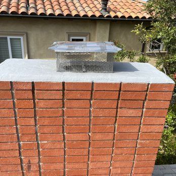 A rectangular brick chimney topped with a gray concrete cap and a metal mesh chimney cover against a house roof.