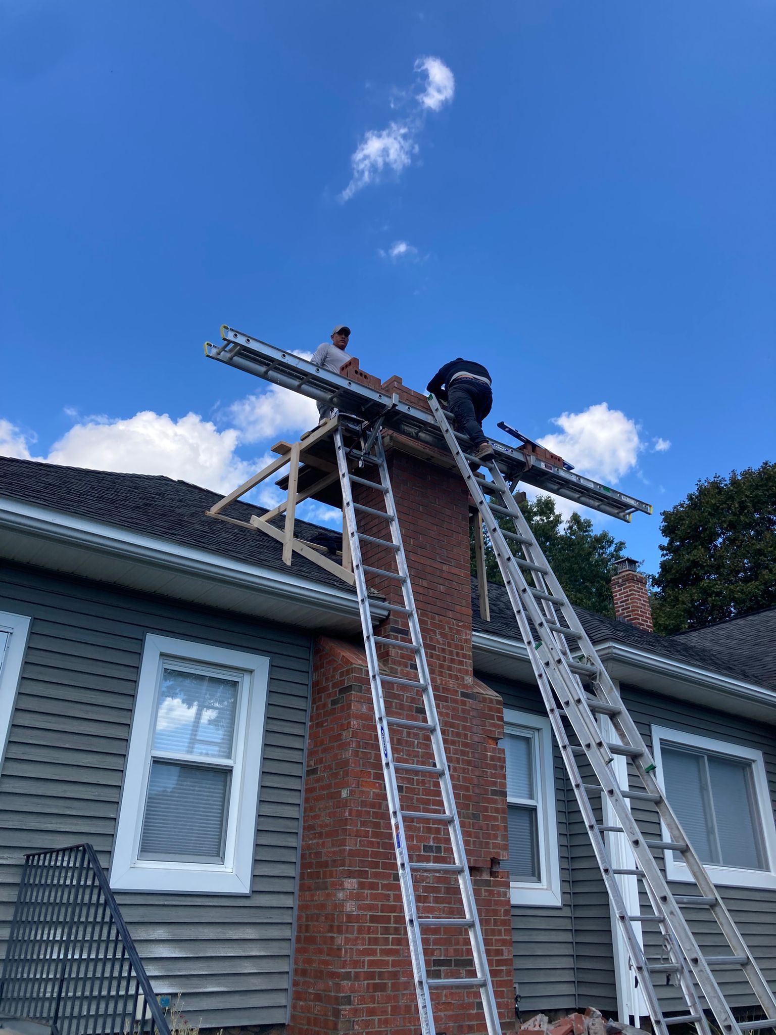 Two people work on a brick chimney repair using ladders and a wooden scaffolding platform on a gray-sided house.