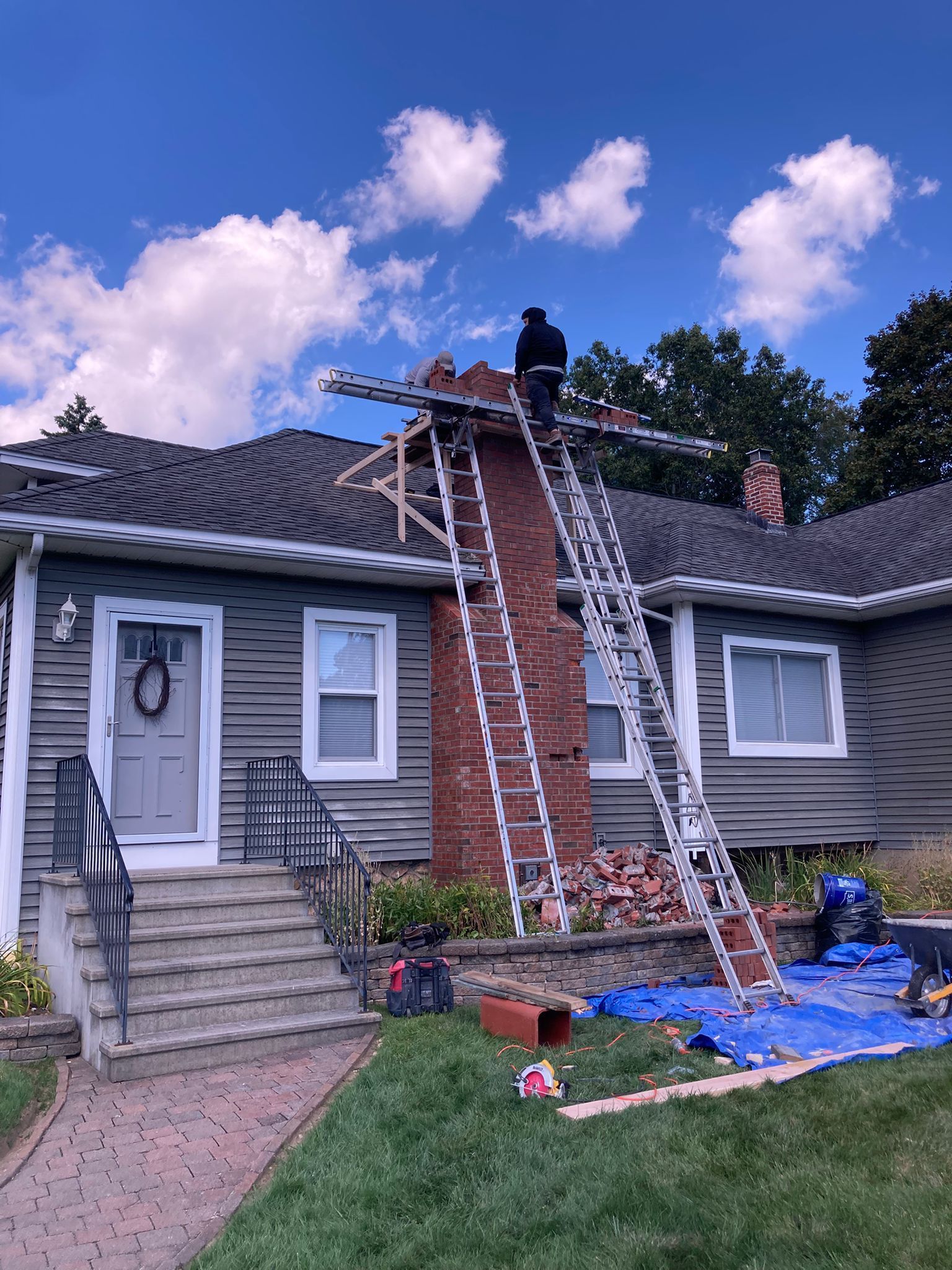 A person on a ladder repairs a brick chimney on the roof of a gray house with a blue tarp spread on the ground below.