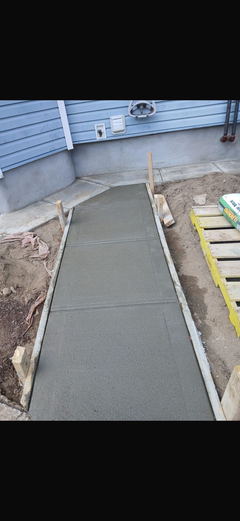 A freshly poured concrete sidewalk is framed by wooden stakes and plastic edging along the side of a blue-sided house.