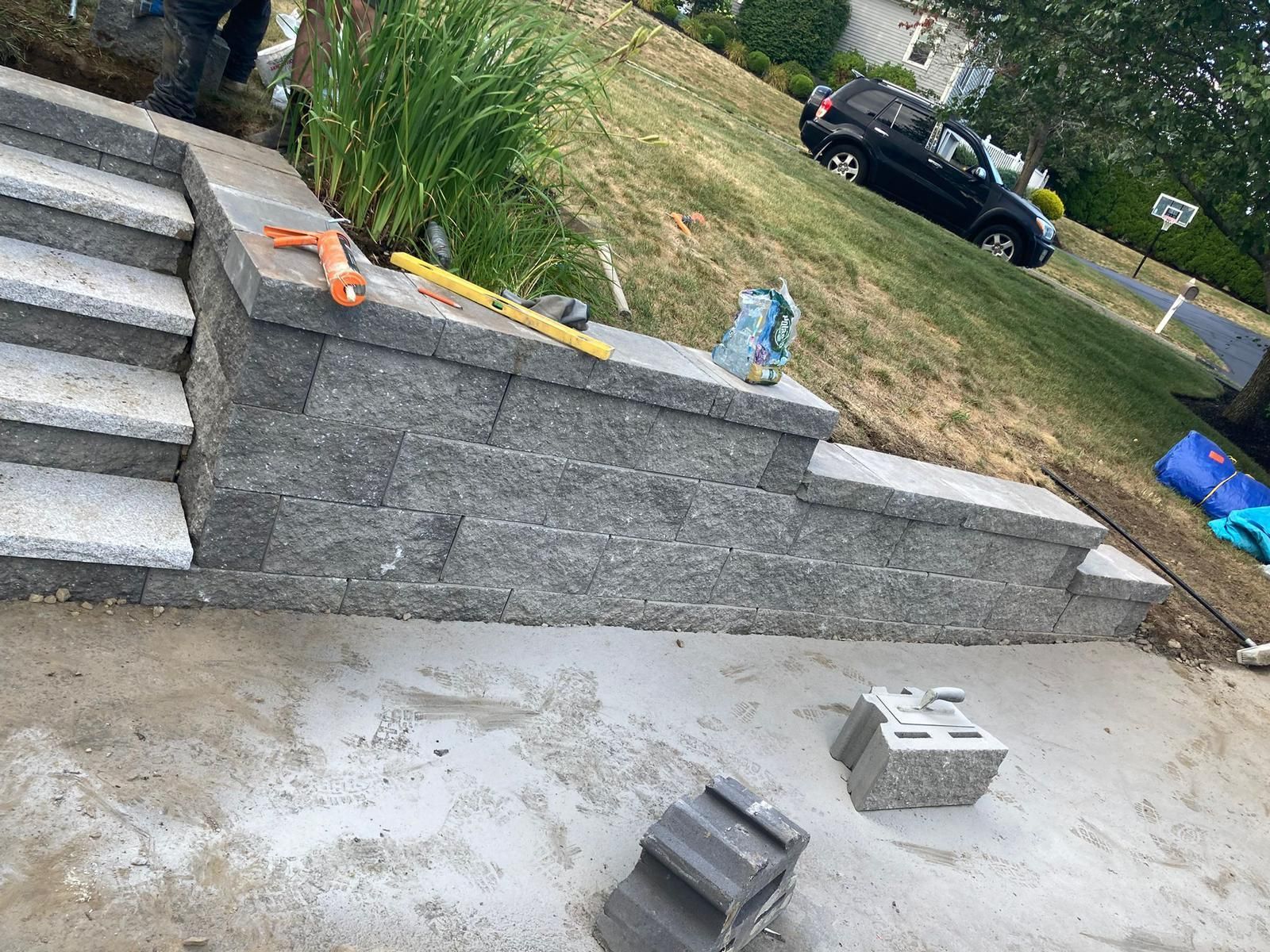 Newly constructed gray stone retaining wall with steps leading up a grassy hill, with tools and construction blocks nearby.