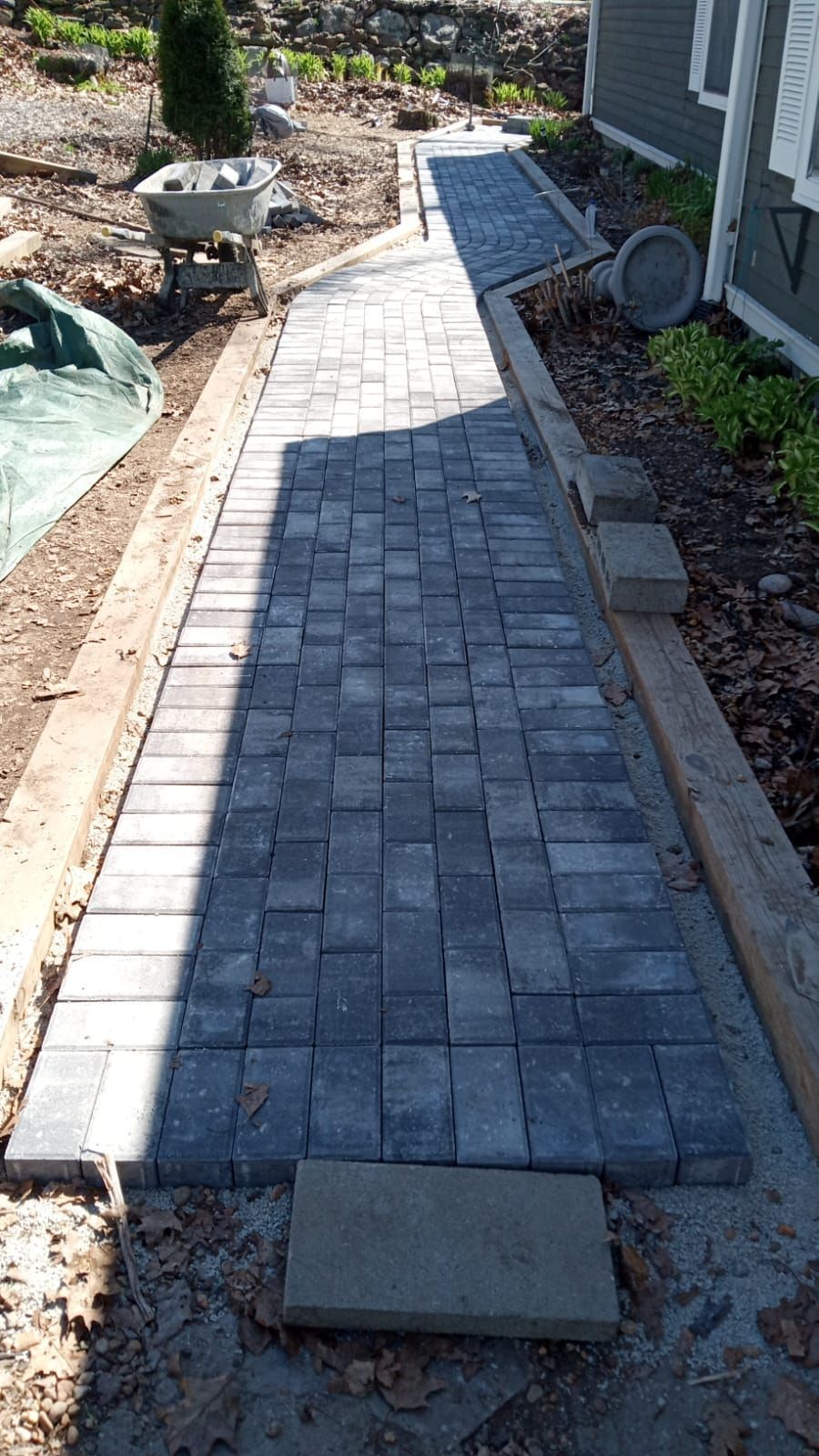 A new gray paver walkway under construction alongside a house, with a border of lighter pavers and surrounding yard debris.