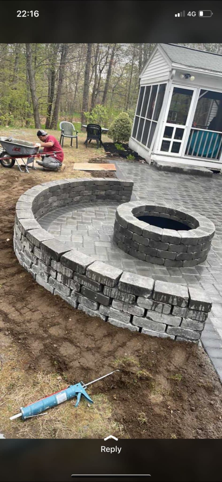 A person in a red shirt works on a patio installation featuring a circular stone fire pit and a curved retaining wall.