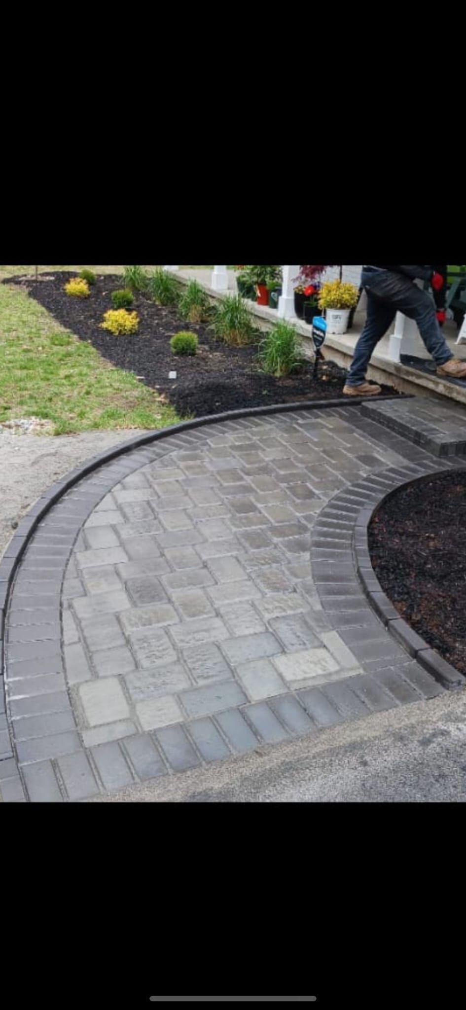 A curved brick paver walkway with a border, near a garden with a person working.