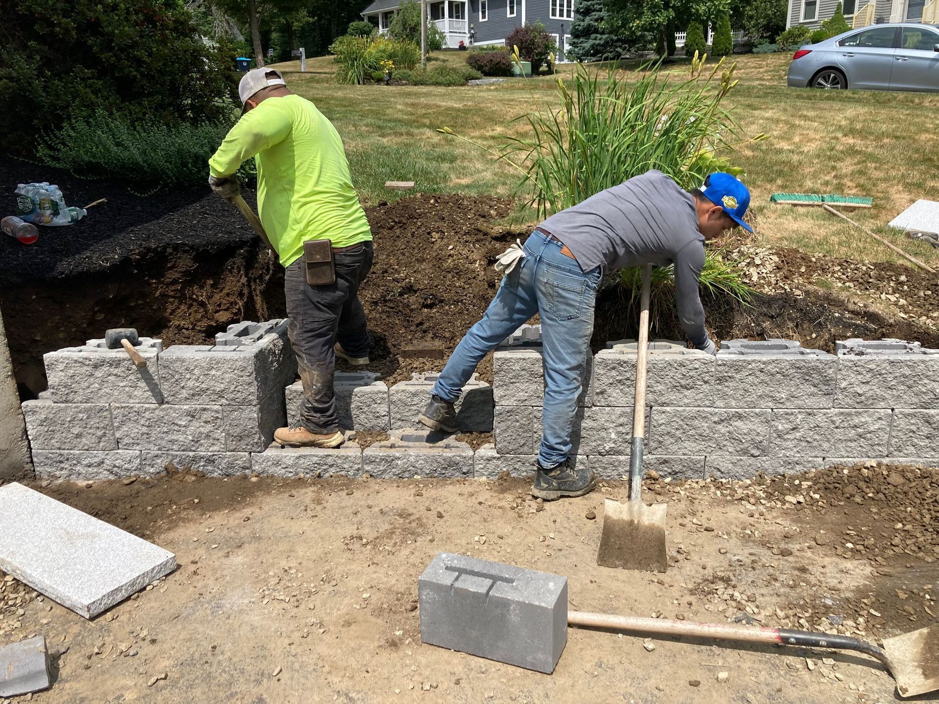 Two workers building a retaining wall with gray blocks outdoors. One shovels soil, the other arranges blocks.