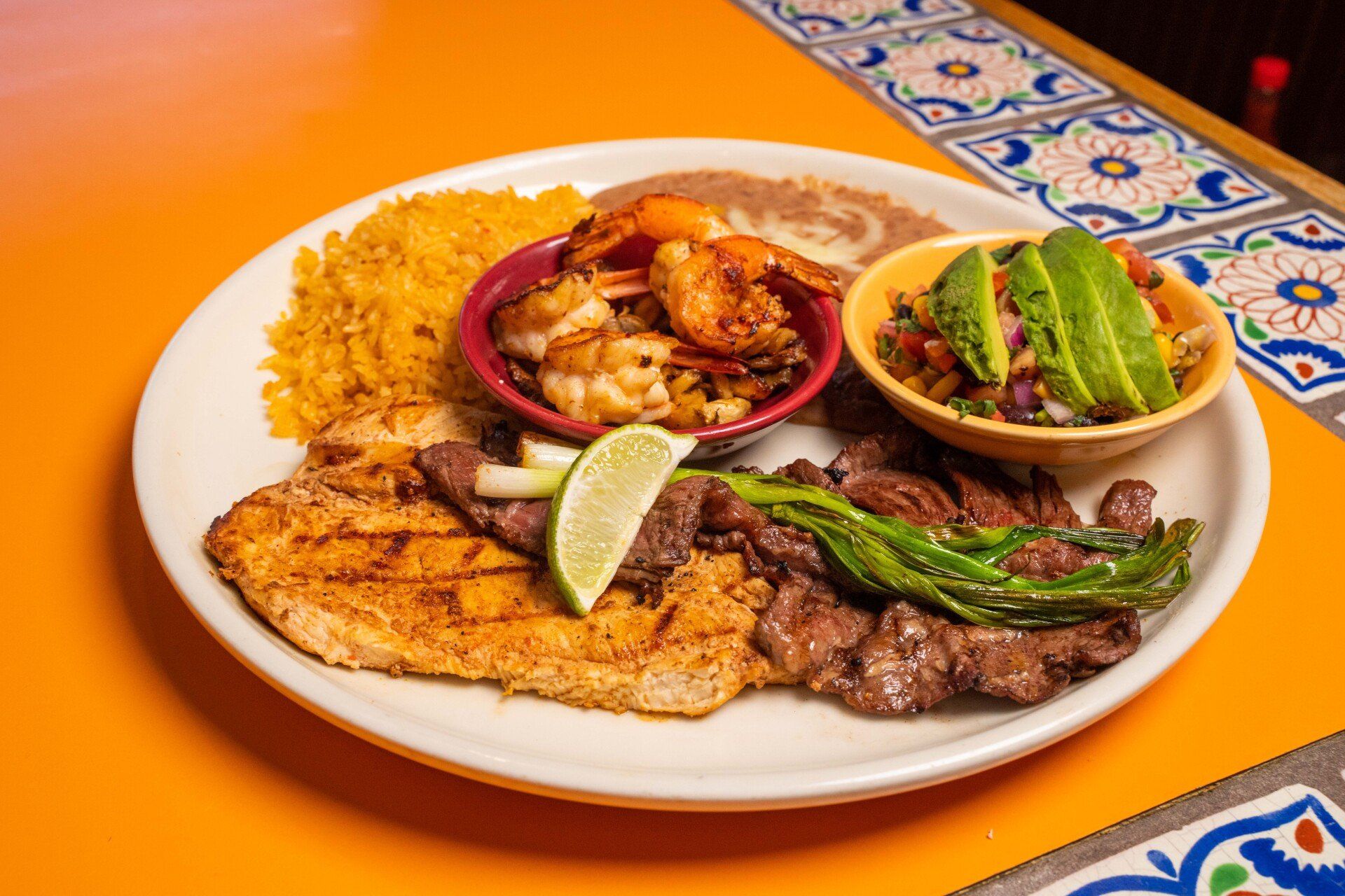 A plate of food with meat, shrimp, rice, and avocado on a table.