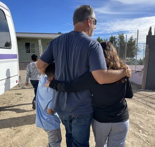A man with his arms around two children, walking towards a building on a sunny day.