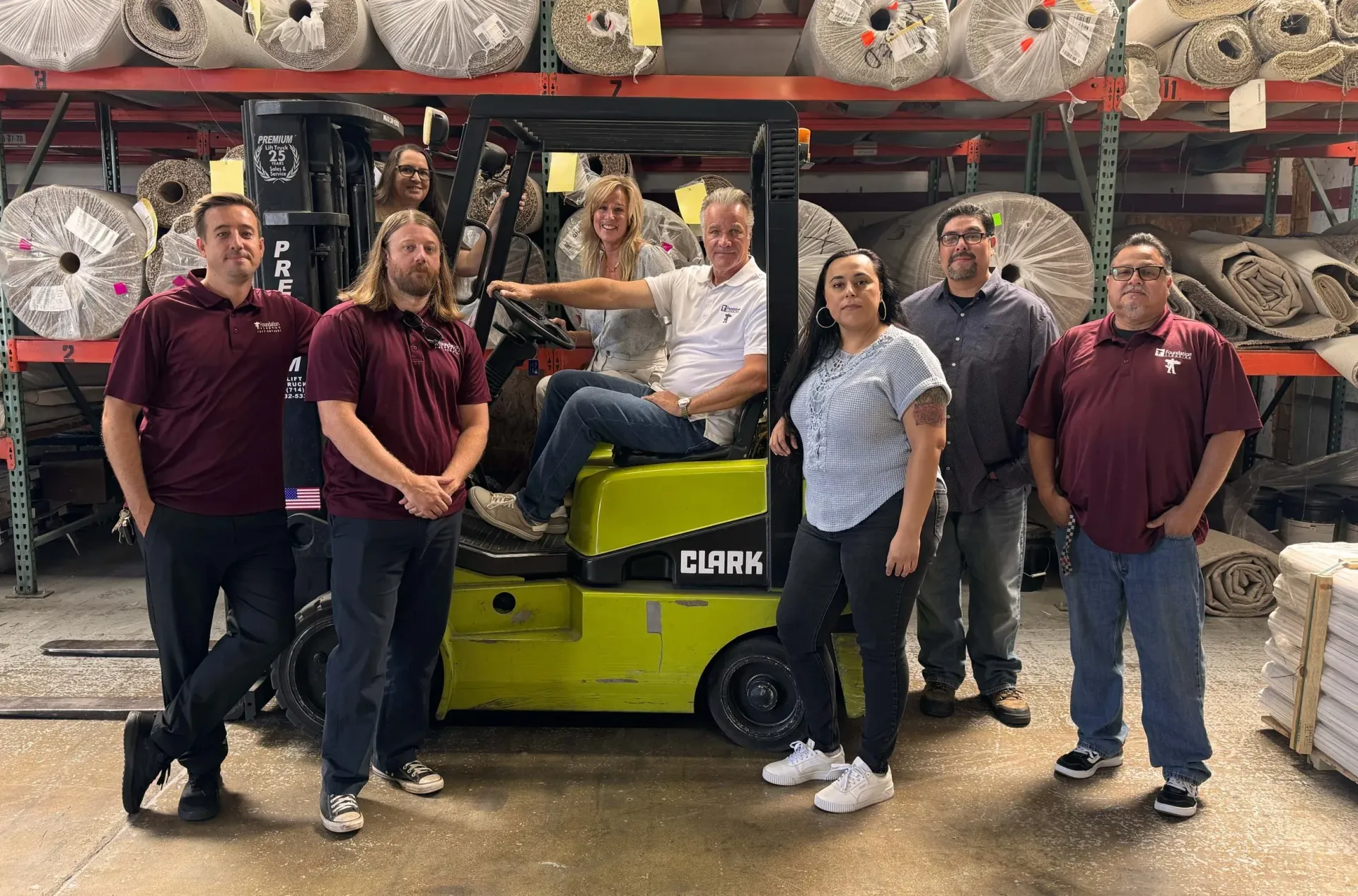 Group of people posing with a forklift in a warehouse filled with carpet rolls. Some are sitting on it.