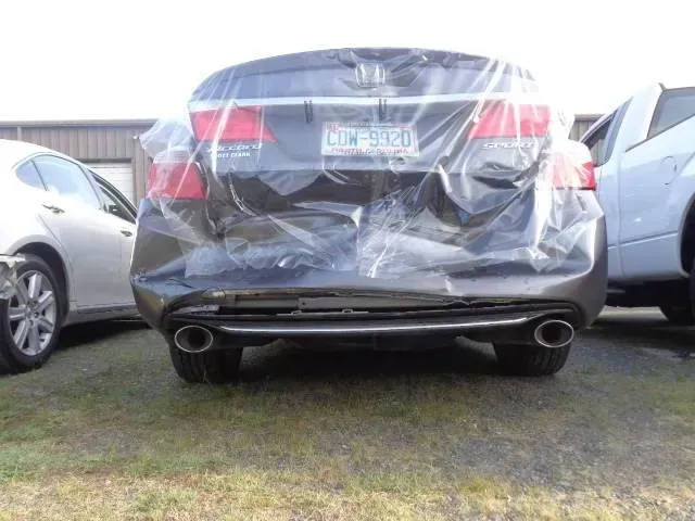 Rear view of a black sedan with plastic covering the trunk, parked between two cars on grass.