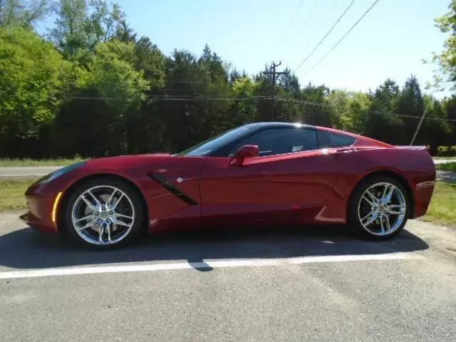 Red sports car parked on a roadside in bright daylight, with trees and blue sky in the background.