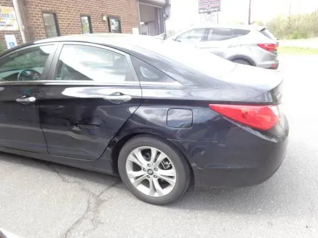 Black sedan parked on a cracked driveway beside a brick house, rear side view