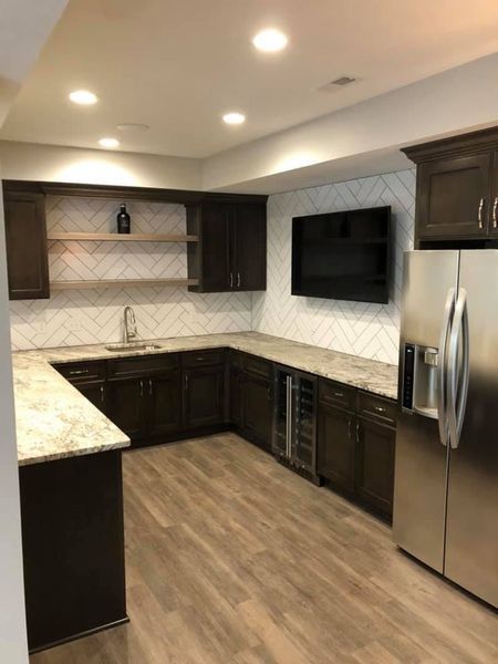 Dark wood bar with granite counters, wine cooler, stainless fridge, and TV on a white and gray tiled wall.