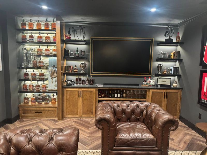 A luxurious home bar with illuminated shelves displaying liquor bottles, a large TV, and leather armchairs.