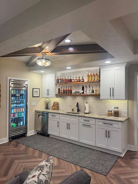 Basement bar with white cabinets, liquor bottles, fridge, and a rug on herringbone floors.