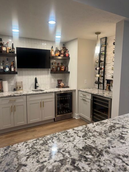 A home bar with white cabinets, granite countertop, shelves with liquor, a wine cooler, and a television.