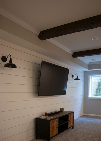TV mounted on shiplap wall, flanked by black sconces. Entertainment console below, beams above.