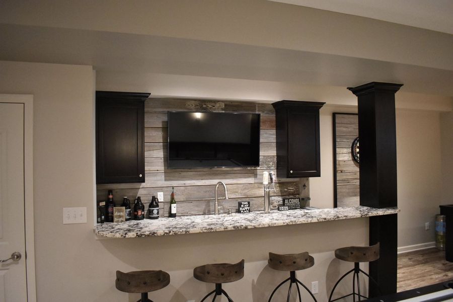 Basement bar with dark cabinets, rustic wood accent wall, granite countertop, stools, and a TV.