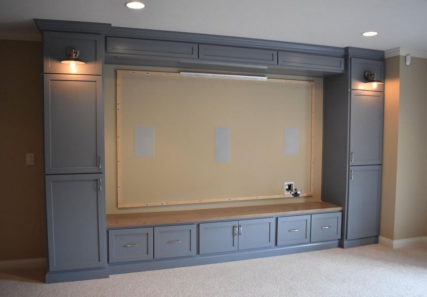 Built-in gray entertainment center with cabinets, drawers, seating, and a blank space for a TV in a room with beige walls and carpet.