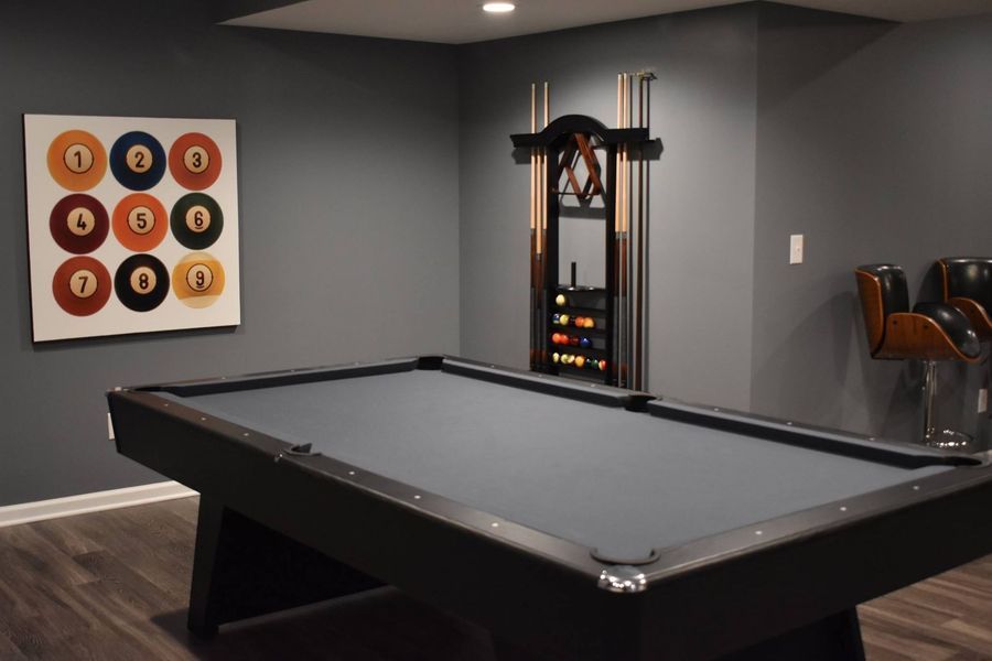 A dark gray pool table in a room with matching walls. Artwork and cue rack are on the wall.