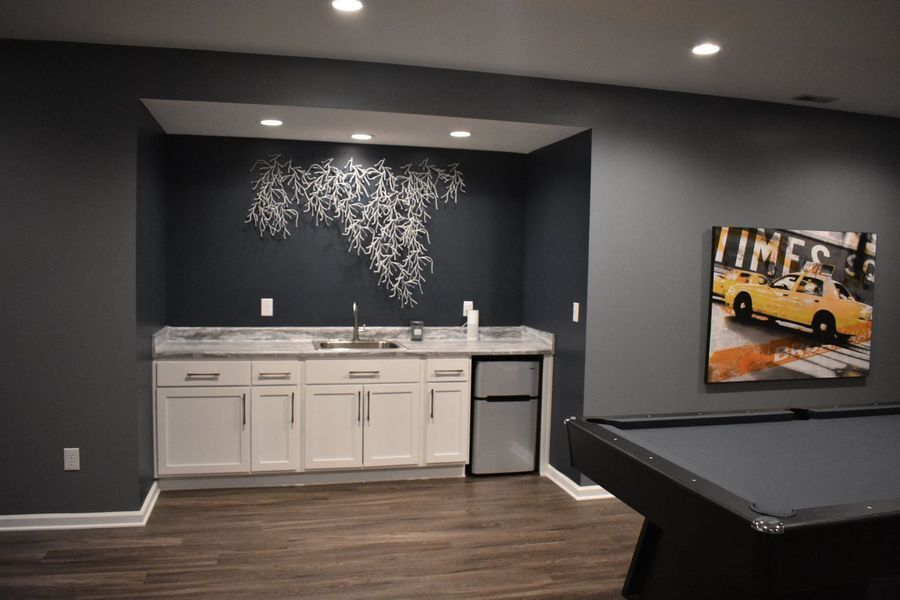 Basement with bar area, pool table. Gray walls, white cabinets, artwork of branches on a dark blue wall.