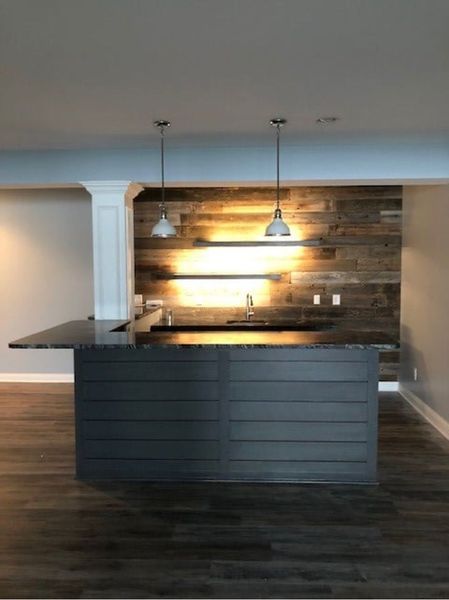 Bar with a dark countertop and paneled grey base. Wood accent wall with sink and pendant lights.
