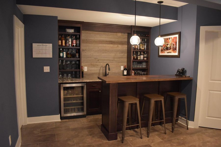 Basement bar with wooden counter, stools, and built-in shelving, lit by pendant lights, dark blue walls.