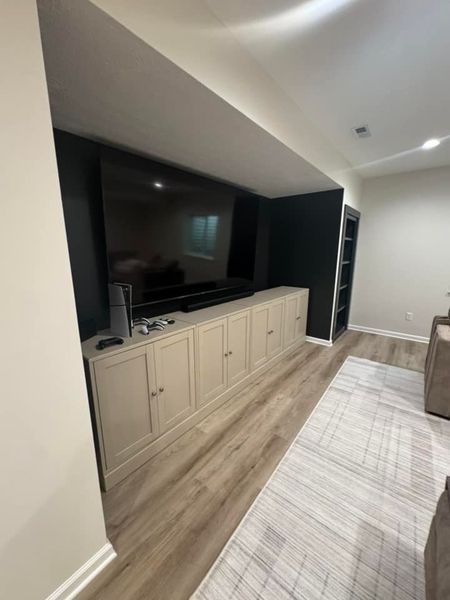 Media room with large TV over built-in storage cabinets. Light wood flooring, gray rug, black accent wall.