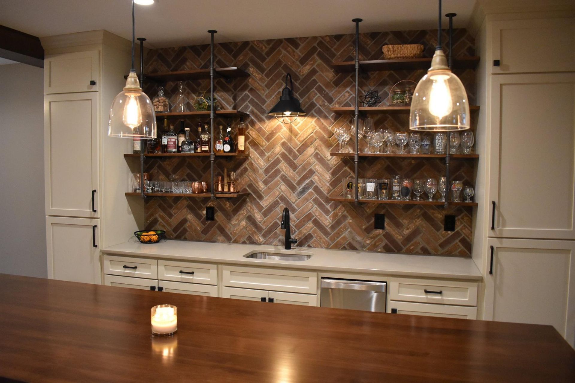 Bar area with wooden shelves, sink, liquor, and glassware against a brown herringbone tiled wall, with pendant lights.
