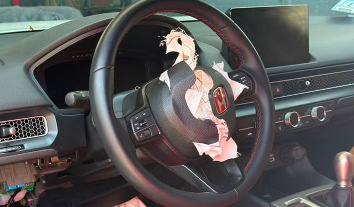 Car interior with black steering wheel and dashboard; a damaged airbag hangs from the center