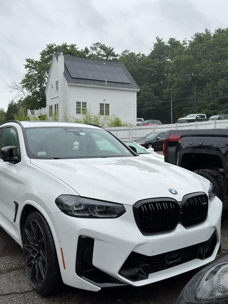 White BMW SUV parked outdoors beside other vehicles and a white house on a cloudy day